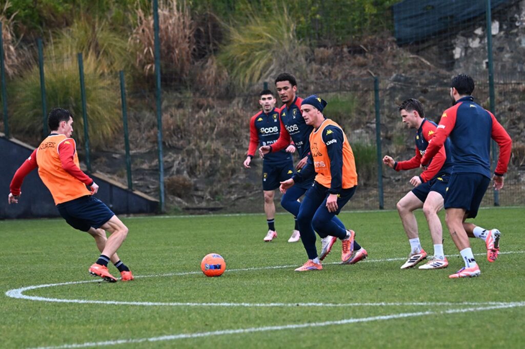Durante l'allenamento del Genoa, i giocatori si esercitano sul campo con pettorine arancioni e maglie rossoblù sotto lo sguardo di De Rossi.