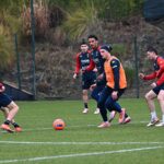 Durante l'allenamento del Genoa, i giocatori si esercitano sul campo con pettorine arancioni e maglie rossoblù sotto lo sguardo di De Rossi.