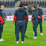 Alcuni calciatori del Genoa in tuta blu scuro con inserti rossi si radunano sul prato dello stadio Olimpico di Roma durante il riscaldamento prepartita.