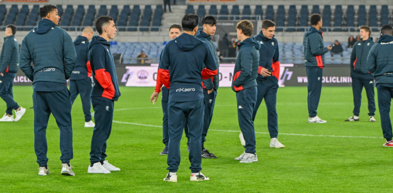 Alcuni calciatori del Genoa in tuta blu scuro con inserti rossi si radunano sul prato dello stadio Olimpico di Roma durante il riscaldamento prepartita.
