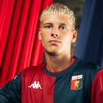 Albert Gronbaek posa con la maglia del Genoa a strisce rossoblù, davanti a tende rosse e blu, nel contesto del passaggio ufficiale all’Amburgo.