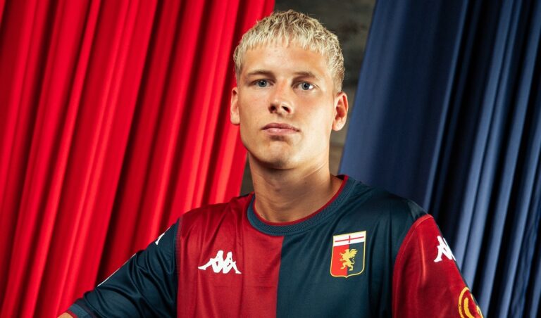 Albert Gronbaek posa con la maglia del Genoa a strisce rossoblù, davanti a tende rosse e blu, nel contesto del passaggio ufficiale all’Amburgo.