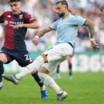 Durante Lazio-Genoa due calciatori si contendono un pallone in scivolata all'Olimpico, con maglie biancocelesti e rossoblù sotto gli spalti.