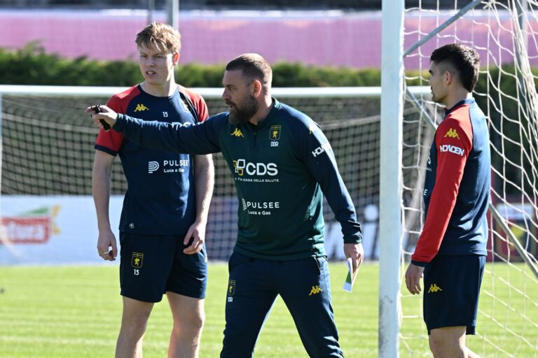 Durante l’allenamento del Genoa, Daniele De Rossi indica la posizione ai giocatori in campo, con maglie blu e rosse vicino alla porta del centro sportivo.