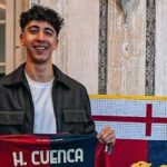 Sorridente, Hugo Cuenca tiene in mano una maglia rossoblù con il nome H. Cuenca, davanti a un pannello con croce rossa e blu in interno.