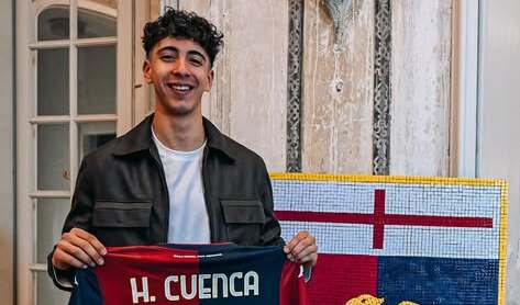 Sorridente, Hugo Cuenca tiene in mano una maglia rossoblù con il nome H. Cuenca, davanti a un pannello con croce rossa e blu in interno.