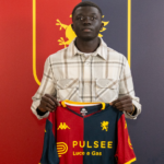 Chec Bebel Doumbia tiene in mano la maglia del Genoa davanti a uno sfondo rosso e blu, con lo sguardo rivolto verso la fotocamera.