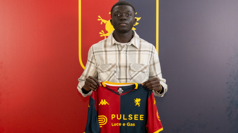 Chec Bebel Doumbia tiene in mano la maglia del Genoa davanti a uno sfondo rosso e blu, con lo sguardo rivolto verso la fotocamera.