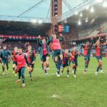 Il Genoa festeggia sul campo a fine partita, con i giocatori in maglia rossoblù che saltano e alzano le braccia nello stadio gremito.
