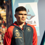 Un giocatore del Genoa in tuta rosso-blu con patch Comune di Sanremo aspetta l'ingresso in campo davanti al logo della Serie A.