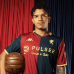 Un giovane calciatore del Genoa posa in maglia rossoblù con sponsor Pulsee Luce e Gas, davanti a tende rosse e blu, con un vecchio pallone sotto il braccio.