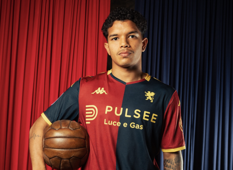 Un giovane calciatore del Genoa posa in maglia rossoblù con sponsor Pulsee Luce e Gas, davanti a tende rosse e blu, con un vecchio pallone sotto il braccio.