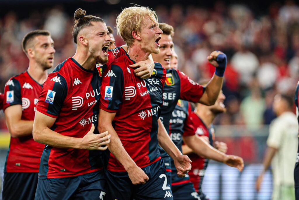 Morten Thorsby esulta con i compagni del Genoa in maglia rossoblù dopo una giocata decisiva, mentre lo stadio rumoreggia sullo sfondo.