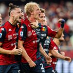 Morten Thorsby esulta con i compagni del Genoa in maglia rossoblù dopo una giocata decisiva, mentre lo stadio rumoreggia sullo sfondo.