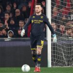 Bijlow, portiere del Genoa, in tenuta scura con numero 16 e guanti chiari, sul prato davanti alla porta mentre osserva il pallone, tra tifosi in tribuna.