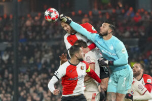 Bijlow, portiere del Genoa, in volo tra avversari durante un contrasto aereo; portiere alto, presenza attiva, lettura dello spazio e gioco con i piedi.