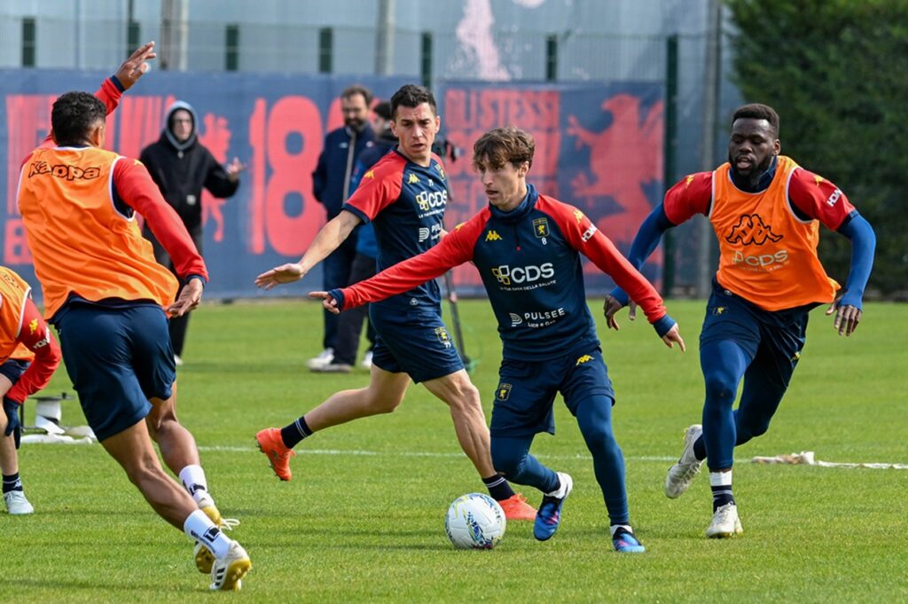 Genoa: giocatori in kit blu e casacche arancioni durante allenamento, primo piano su atleta con palla, sfondo campo e pannelli rossi, maschile concentrato, dettagli distintivi CDS