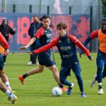 Genoa: giocatori in kit blu e casacche arancioni durante allenamento, primo piano su atleta con palla, sfondo campo e pannelli rossi, maschile concentrato, dettagli distintivi CDS