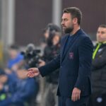 De Rossi in giacca blu con stemma Genoa, barba curata e mano in gesto tecnico durante la partita di Roma allo stadio Luigi Ferraris.