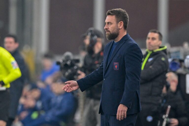De Rossi in giacca blu con stemma Genoa, barba curata e mano in gesto tecnico durante la partita di Roma allo stadio Luigi Ferraris.
