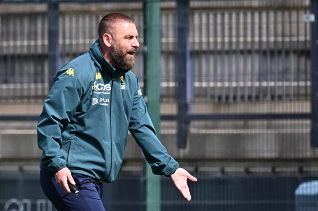 De Rossi, allenatore del Genoa, ritratto durante l’allenamento in giacca sportiva blu scuro, gesto energico con una mano aperta, sfondo recinzione sportiva e campo.