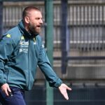 De Rossi, allenatore del Genoa, ritratto durante l’allenamento in giacca sportiva blu scuro, gesto energico con una mano aperta, sfondo recinzione sportiva e campo.