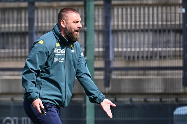 De Rossi, allenatore del Genoa, ritratto durante l’allenamento in giacca sportiva blu scuro, gesto energico con una mano aperta, sfondo recinzione sportiva e campo.