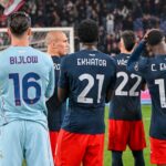 Bijlow tra i giocatori in maglie blu scuro e shorts rossi, Ostigard e Ellertsson tra i compagni in primo piano in un stadio gremito, con testo calcistico sullo sfondo