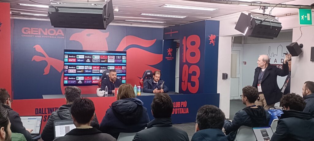 Sala stampa Genoa con pannelli rosso e blu e scritta 1893: De Rossi parla ai giornalisti mentre Patrizio Masini segue i lavori