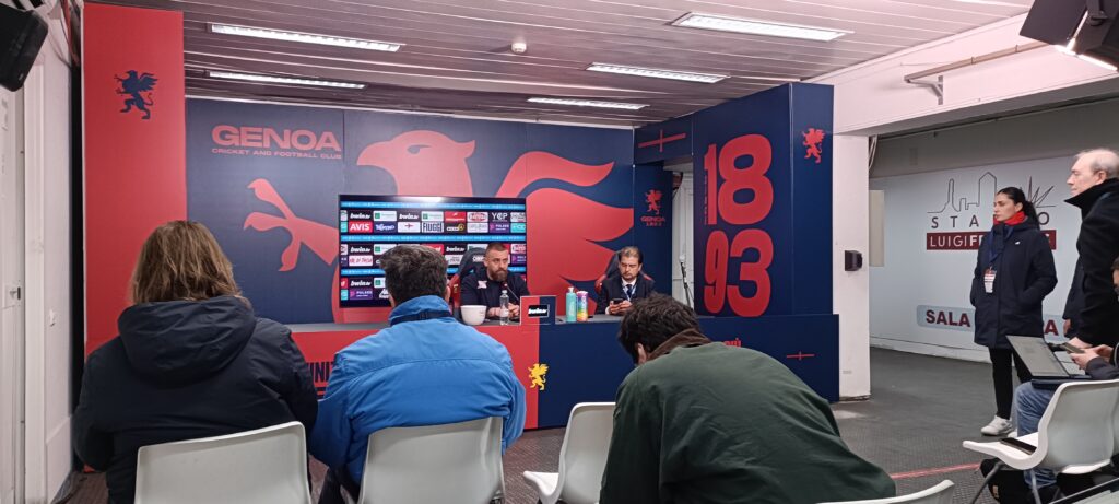 Conferenza stampa Genoa: Daniele De Rossi al tavolo con altri membri, sfondo blu con leone rosso e logo Genoa, numeri 18 e 93, bottiglie colorate sul tavolo.