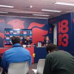 Conferenza stampa Genoa: Daniele De Rossi al tavolo con altri membri, sfondo blu con leone rosso e logo Genoa, numeri 18 e 93, bottiglie colorate sul tavolo.