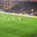 Messias in azione su campo verde in uno stadio gremito: riflessi gialli delle pubblicità e atmosfera da Ferraris con i colori rossoblù.