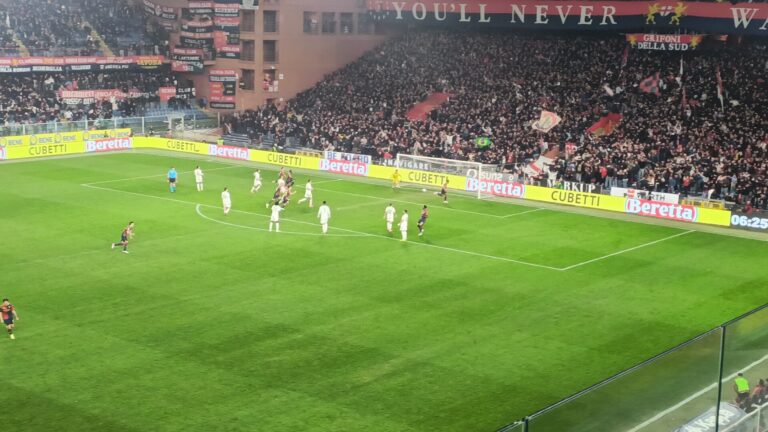 Messias in azione su campo verde in uno stadio gremito: riflessi gialli delle pubblicità e atmosfera da Ferraris con i colori rossoblù.