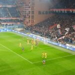 Malinovskyi tra i giocatori del Genoa in maglia rossoblu, tifosi in curva con striscioni, azione di calcio al stadio tra Genoa e Udinese durante una partita intensa