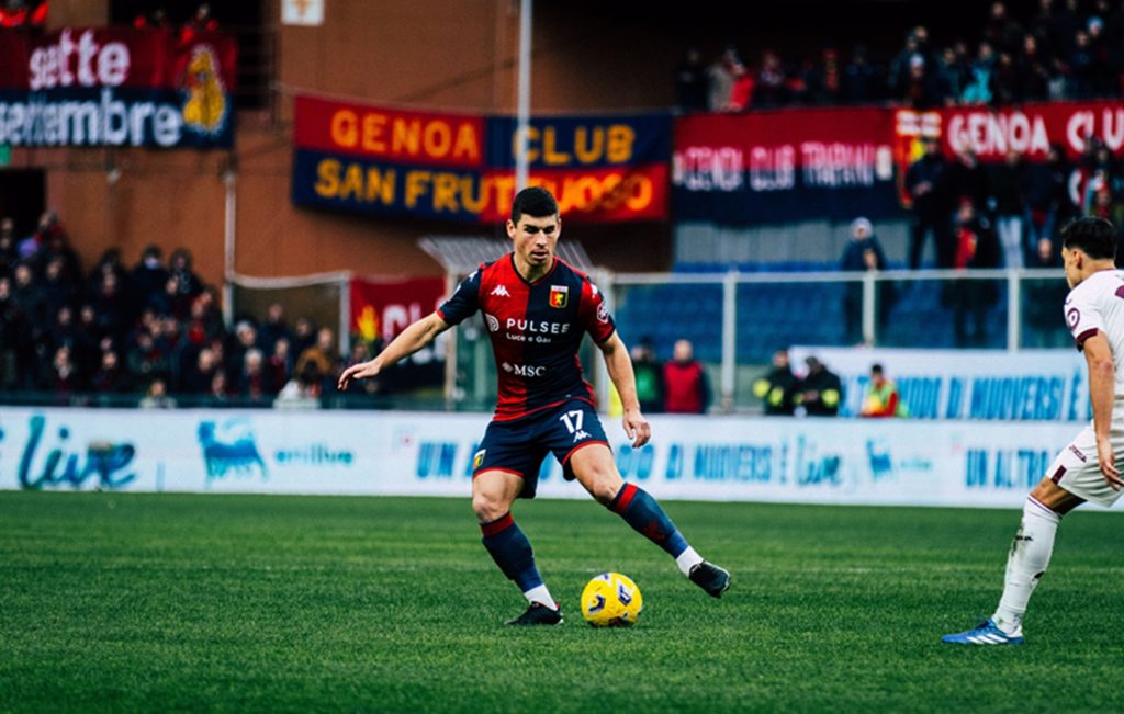 Malinosvkyi del Genoa in maglia rossoblù sul prato verde al Bentegodi, mentre controlla il pallone giallo. Verona-Genoa, finale salvezza.