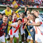 Genoa e Verona celebrano, giocatori in divise bianco-rosse e gialle, volti sorridenti, braccia alzate in festa sul prato, tifosi sugli spalti alimentano l'energia della scena