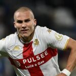 Leo Østigård, difensore rossoblù, in azione sul campo con la maglia bianca e rossa del Genoa, esprime détermination e dinamismo, insieme a riferimenti a Vitinha e Dimarco