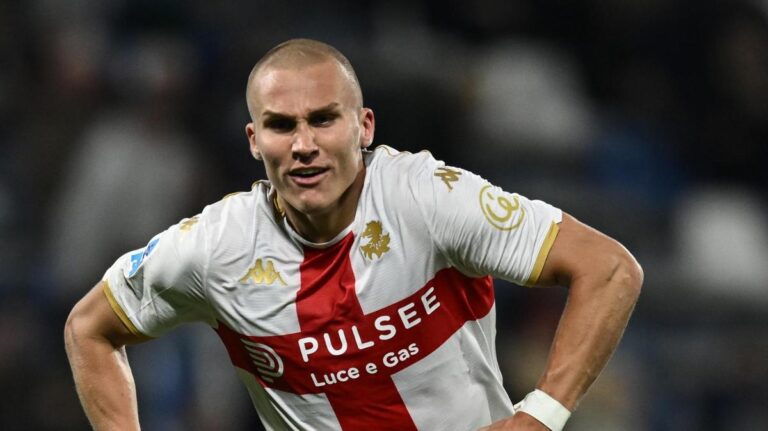 Leo Østigård, difensore rossoblù, in azione sul campo con la maglia bianca e rossa del Genoa, esprime détermination e dinamismo, insieme a riferimenti a Vitinha e Dimarco
