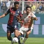 Nel derby Genoa-Roma, De Rossi segue la lotta in campo: giocatori con maglie blu-rosse e bianco-rosse contendono il pallone.