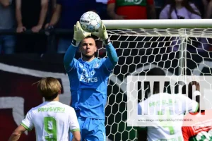 Portiere in completo blu solleva la palla da calcio sopra la testa, guanti da portiere, davanti a una rete, pubblico sullo sfondo, richiamo a Bijlow e al Genoa
