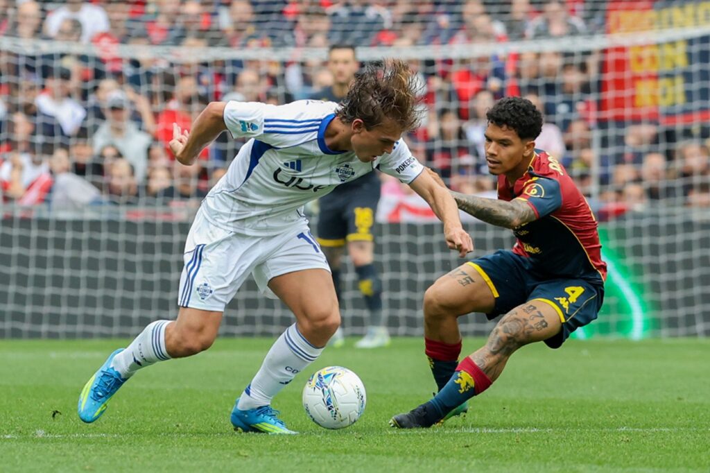 Contrasto in area tra Genoa e Como al Luigi Ferraris: calciatori in maglia bianco-blu contro divisa rosso-nera, palla in primo piano e rete sullo sfondo.