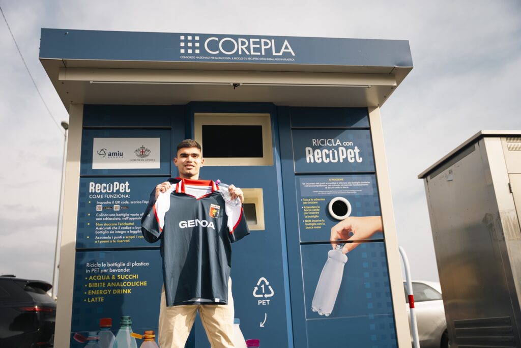 Maglia del Genoa in blu con dettagli rossi, mostrata di fronte a una cabina RECOPET COREPLA, con loghi Genoa e Corepla, rimarcando elementi riciclati in PET e sostenibilità.