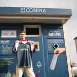 Maglia del Genoa in blu con dettagli rossi, mostrata di fronte a una cabina RECOPET COREPLA, con loghi Genoa e Corepla, rimarcando elementi riciclati in PET e sostenibilità.