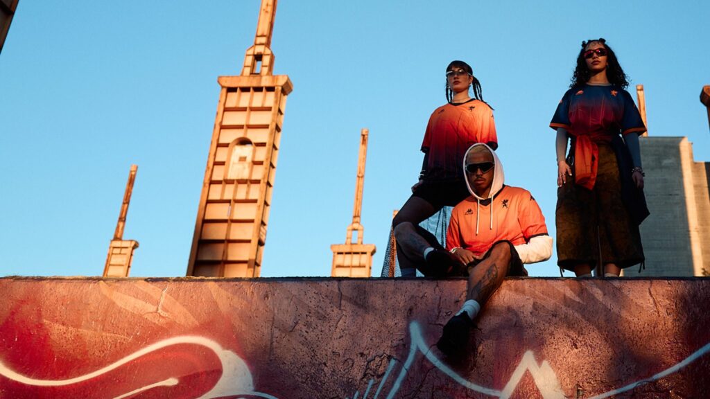 Tre modelli in stile streetwear indossano maglie arancioni con sfumature rosse su un muretto davanti a un cielo azzurro; graffiti in primo piano e strutture urbane sullo sfondo, richiama Genoa CFC e Kappa FuturFestival