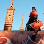 Tre modelli in stile streetwear indossano maglie arancioni con sfumature rosse su un muretto davanti a un cielo azzurro; graffiti in primo piano e strutture urbane sullo sfondo, richiama Genoa CFC e Kappa FuturFestival