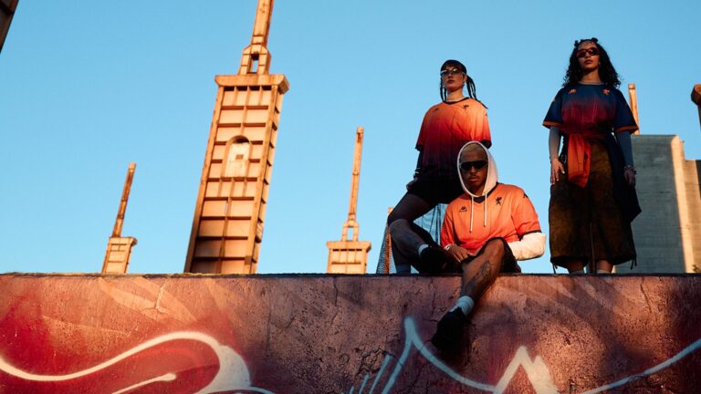 Tre modelli in stile streetwear indossano maglie arancioni con sfumature rosse su un muretto davanti a un cielo azzurro; graffiti in primo piano e strutture urbane sullo sfondo, richiama Genoa CFC e Kappa FuturFestival