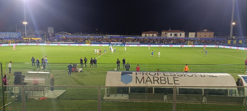 Partita notturna di calcio al campionato tra Spezia e Carrarese, stadio illuminato con tifosi in tribuna, campo verde brillante e panchine laterali