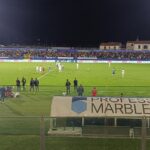 Partita notturna di calcio al campionato tra Spezia e Carrarese, stadio illuminato con tifosi in tribuna, campo verde brillante e panchine laterali