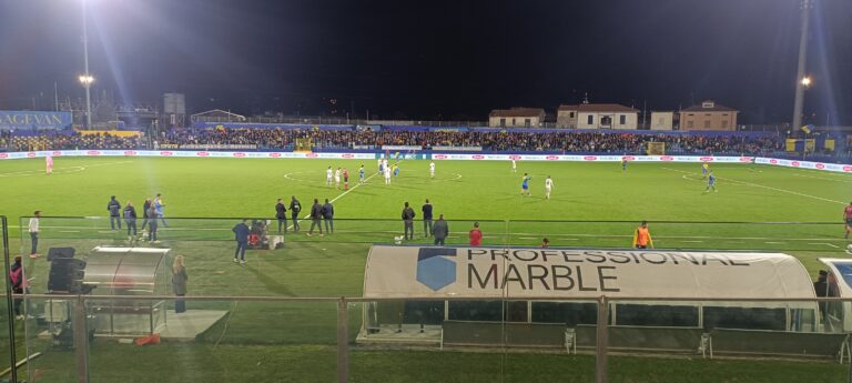 Partita notturna di calcio al campionato tra Spezia e Carrarese, stadio illuminato con tifosi in tribuna, campo verde brillante e panchine laterali