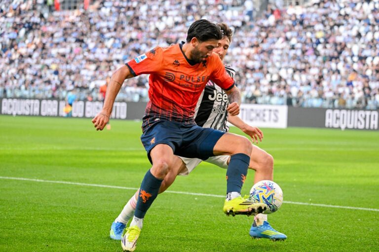Ellertsson in maglia arancione del Genoa sfida un giocatore della Juventus su un prato verde, stando vicino al pallone e con crowd sugli spalti in sfondo